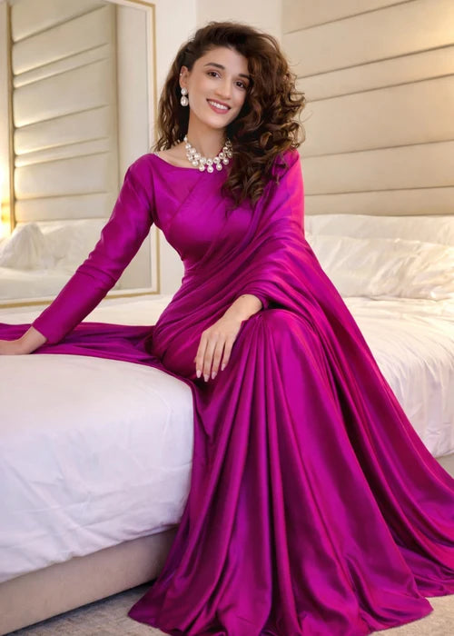 A woman wearing a long-sleeved magenta purple satin gown sits on a white bed in a bedroom setting.