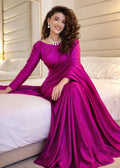 A woman wearing a long-sleeved magenta purple satin gown sits on a white bed in a bedroom setting.