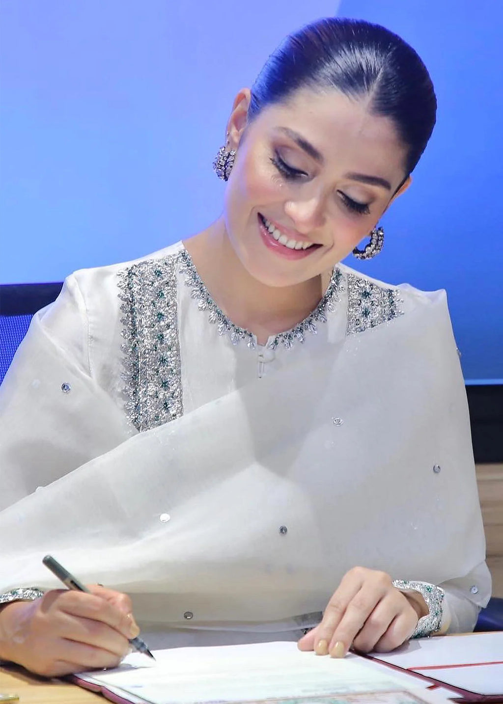 Ayeza khan in a white traditional outfit with silver embroidery is seated at a desk, smiling and writing on a document. She is wearing silver earrings and bracelets against a blue background.