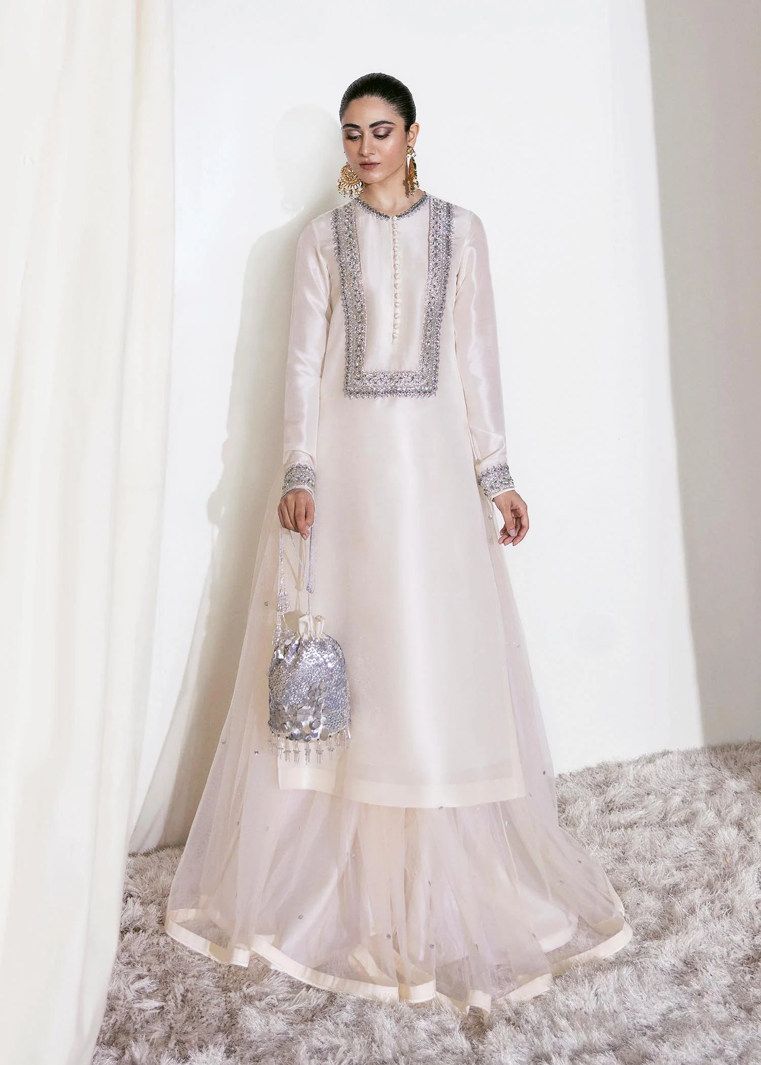 Woman in a white traditional outfit with intricate detailing, holding a decorative bag against a white curtain background.