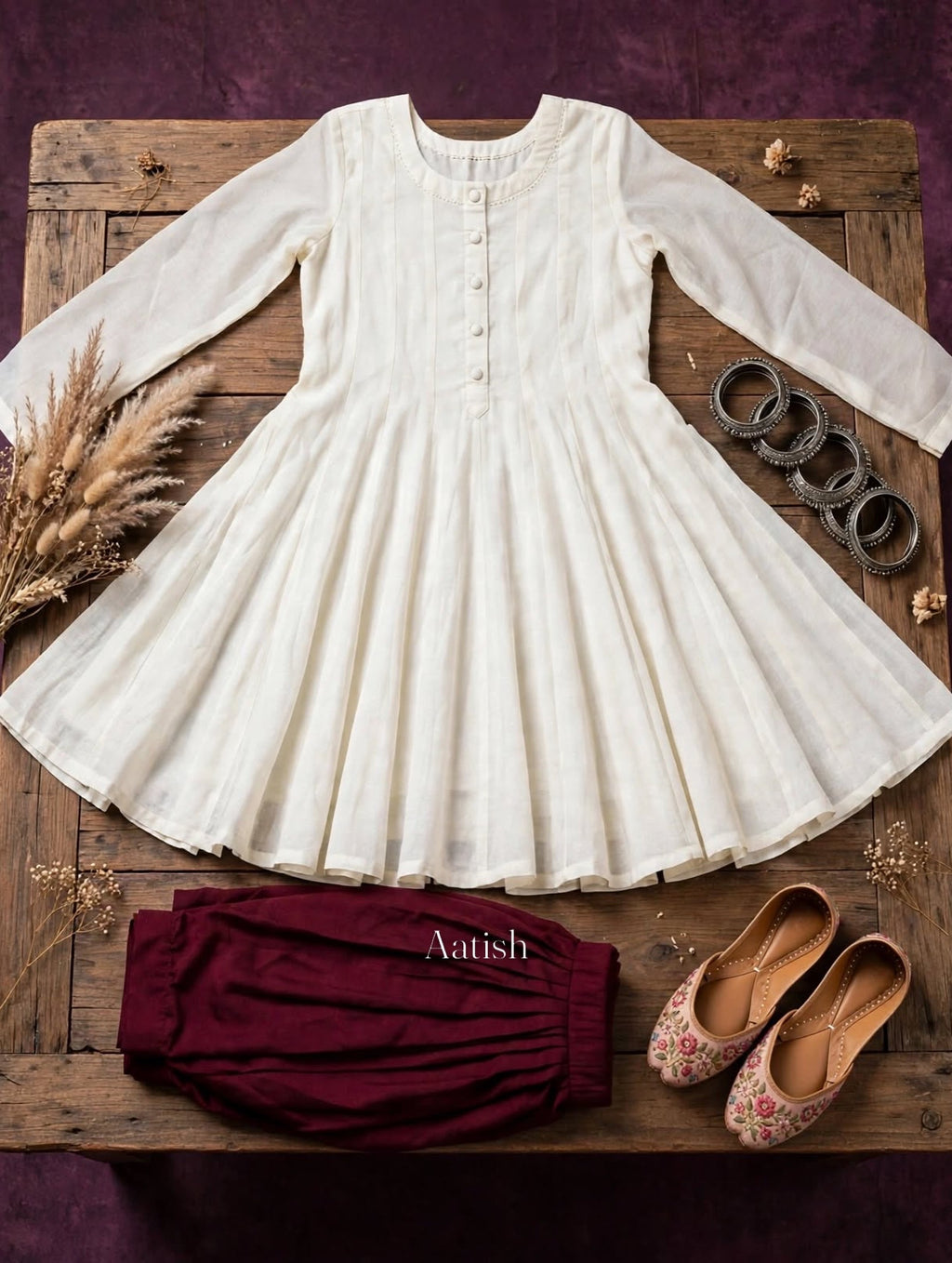 An off-white long-sleeve tunic dress with a buttoned neckline and flared skirt, styled on a rustic wooden board with burgundy bloomers, floral flats, and silver rings against a dark purple background.
