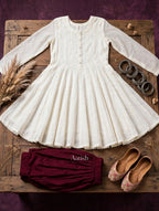 An off-white long-sleeve tunic dress with a buttoned neckline and flared skirt, styled on a rustic wooden board with burgundy bloomers, floral flats, and silver rings against a dark purple background.