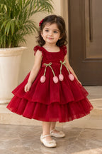 A young girl with dark curly hair wearing a red tiered tulle dress with pink tulip embellishments and white Mary Jane shoes, standing on stone steps in front of a potted plant.