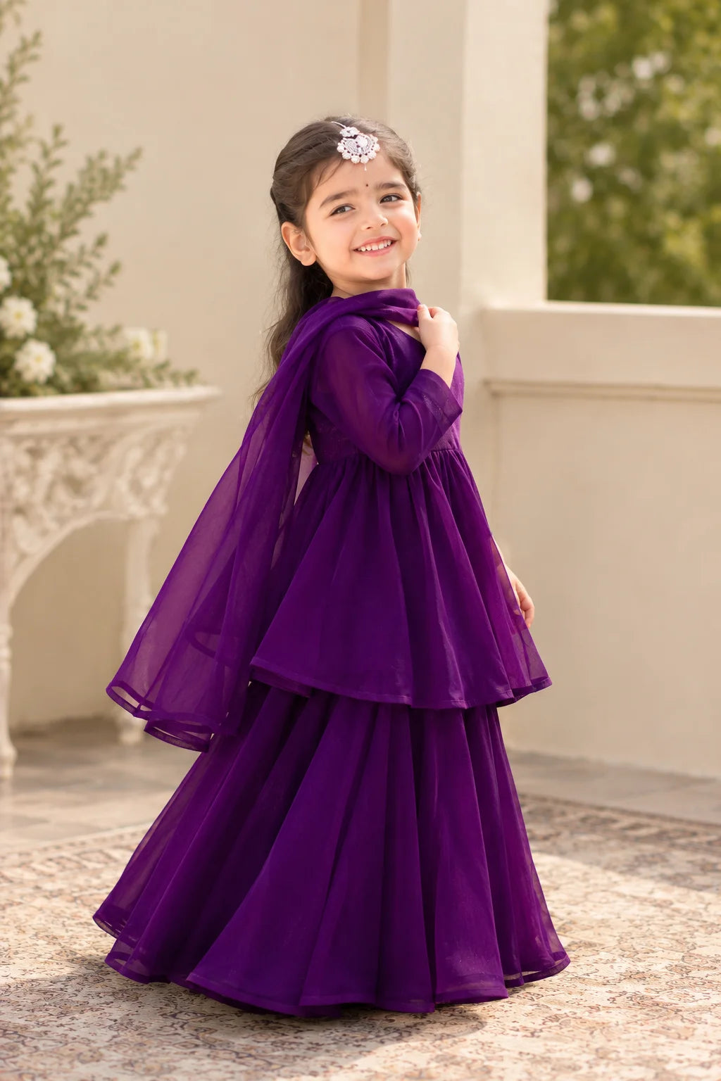 A young girl wearing a purple tiered tunic and flared skirt outfit with a matching sheer dupatta, standing in an outdoor patio setting.