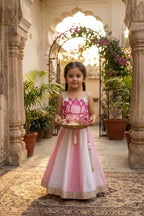 A young girl stands in a sunlit courtyard wearing a pink and white traditional dress with gold embroidery and holding a decorative tray. Stone pillars, potted plants, and an arched walkway are visible in the background.