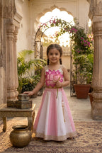 A young girl stands in an ornate courtyard wearing a pink and white traditional dress with a lotus flower motif on the bodice. She holds a lotus flower wand, with stone pillars, potted plants, and a patterned rug in the background.