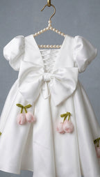 A white satin girls' dress hanging on a pearl-beaded hanger against a gray background. The dress features a large bow at the front, puffed short sleeves, lace-up back detailing, and pink tulip appliqués with green stems.