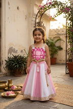 A young girl stands in a sunlit courtyard wearing a pink and white traditional dress with a lotus flower motif on the bodice, gold embroidery, and a full skirt. The setting includes potted plants, a stone floor, and decorative brass vessels.