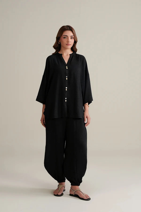 A woman modeling a black two-piece outfit consisting of a loose, button-front long-sleeve top and matching wide-leg pants, paired with brown sandals, against a plain light beige studio background.