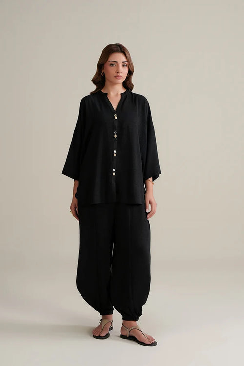 A woman modeling a black two-piece outfit consisting of a loose, button-front long-sleeve top and matching wide-leg pants, paired with brown sandals, against a plain light beige studio background.