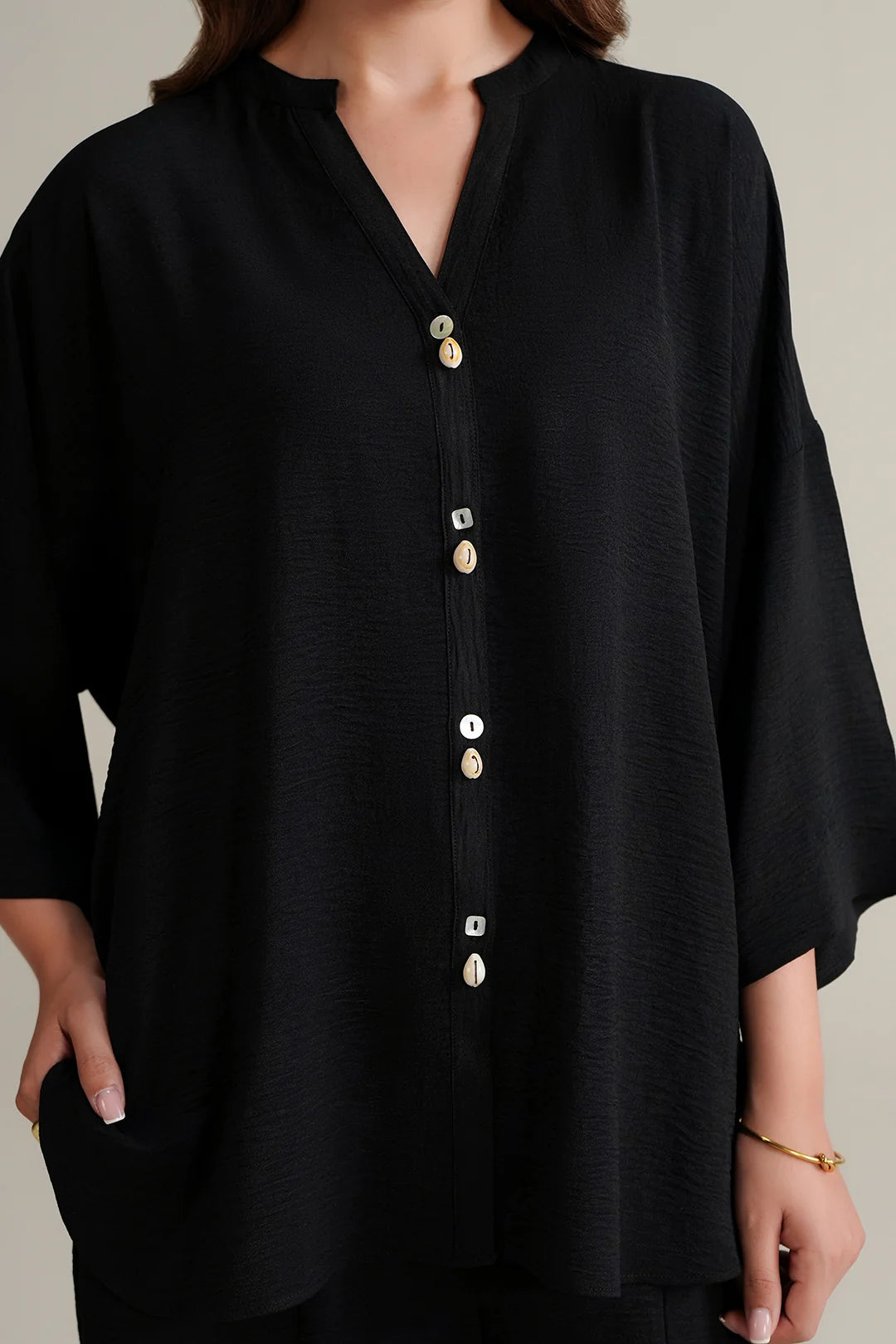 A close-up of a woman wearing a black textured shirt with a split neckline, wide three-quarter sleeves, and decorative gold buttons down the front.