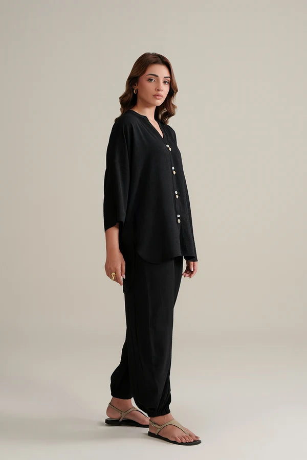 A woman modeling a black two-piece Balloon pant set consisting of a long-sleeve button-front top and matching wide-leg pants, paired with flat sandals, against a solid light beige studio background.