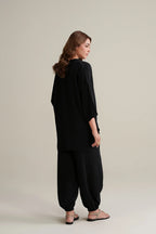 A woman is shown from the back wearing a loose black tunic and matching wide-leg pants, paired with flat sandals, against a solid light beige studio background.