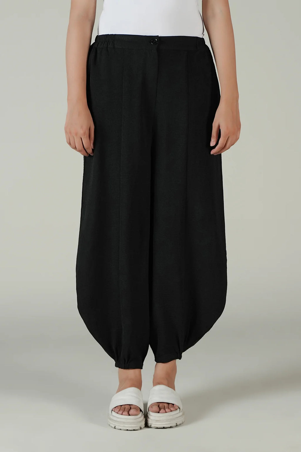 A person wearing loose black balloon-style pants with a white top and white slide sandals, standing against a plain light gray studio background.