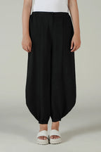 A person wearing loose black balloon-style pants with a white top and white slide sandals, standing against a plain light gray studio background.