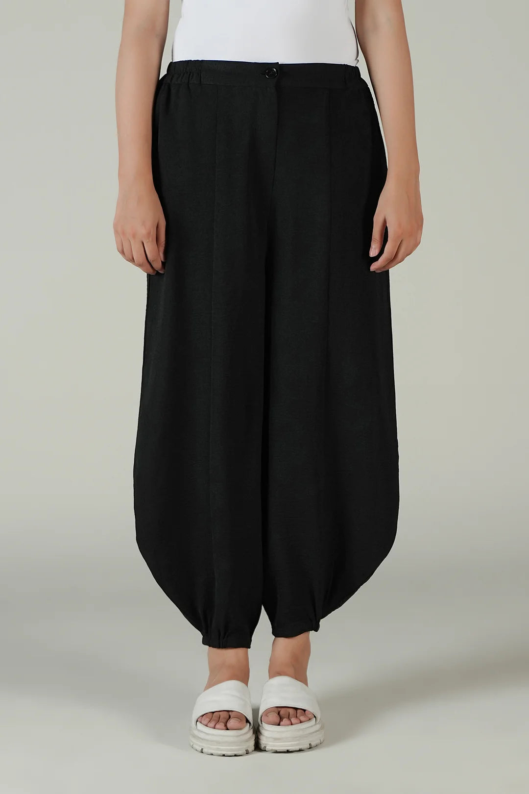 A person wearing loose black balloon-style pants with a white top and white slide sandals, standing against a plain light gray studio background.