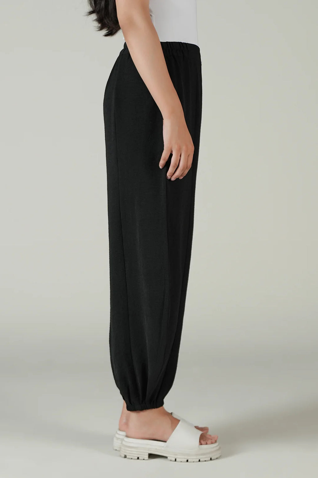 A side view of a person wearing loose black harem-style pants with an elastic waist and gathered cuffs, paired with white slide sandals against a plain light gray studio background.