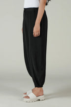 A model wearing black harem-style pants with a gathered elastic waist and ankle cuffs, paired with white slide sandals against a light gray studio background.