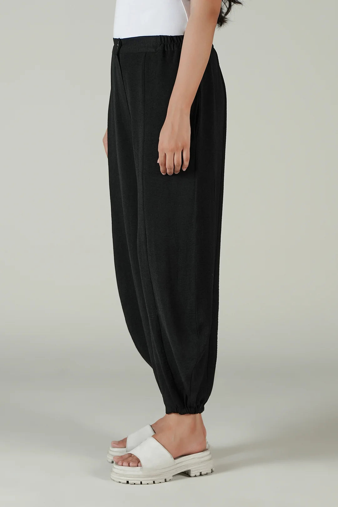 A model wearing black harem-style pants with a gathered elastic waist and ankle cuffs, paired with white slide sandals against a light gray studio background.