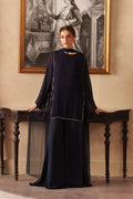 A woman stands indoors wearing a long, dark navy chiffon dress with sheer draped sleeves and a round neckline with plazo and chiffon dupatta. The dress has subtle silver embellishment along the front.