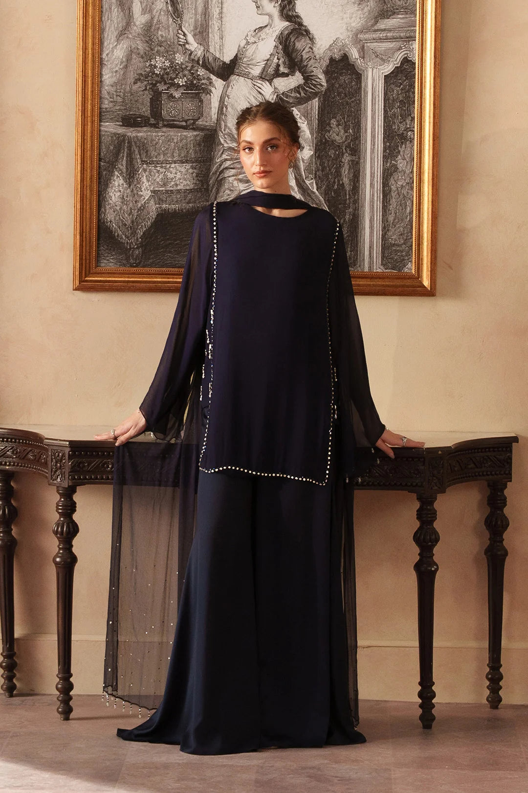 A woman stands indoors wearing a long, dark navy chiffon dress with sheer draped sleeves and a round neckline with plazo and chiffon dupatta. The dress has subtle silver embellishment along the front.