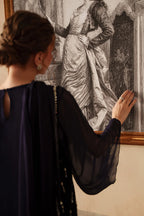 A woman in a dark navy blue sheer-sleeved dress stands in front of a large framed black-and-white classical painting, touching the glass or wall area with her hand.