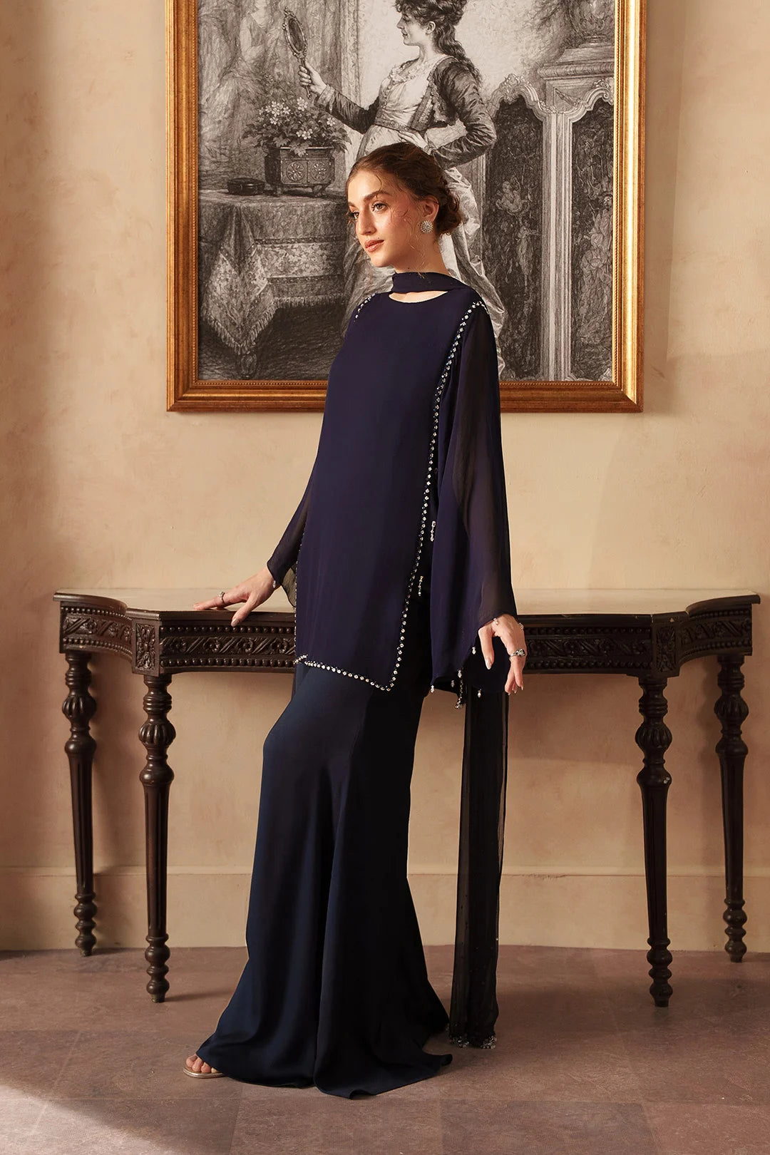A woman models a navy blue three-piece outfit consisting of a long-sleeve top with silver bead trim and matching wide-leg trousers, posed in front of a dark wooden console table and a framed black-and-white painting.