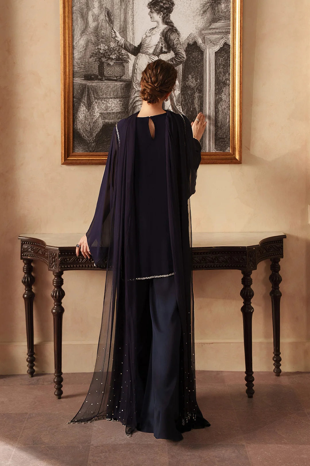 A woman stands with her back to the camera wearing a dark navy blue traditional outfit with a sheer draped overlay and silver embellishments, posed in front of a console table and a framed black-and-white painting in a warm-toned room.