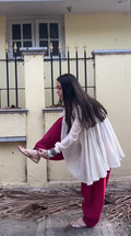 A woman wearing a white long-sleeve tunic and deep red pants stands outdoors in front of a beige wall and black metal fence, holding one foot to show off a pair of flat sandals.