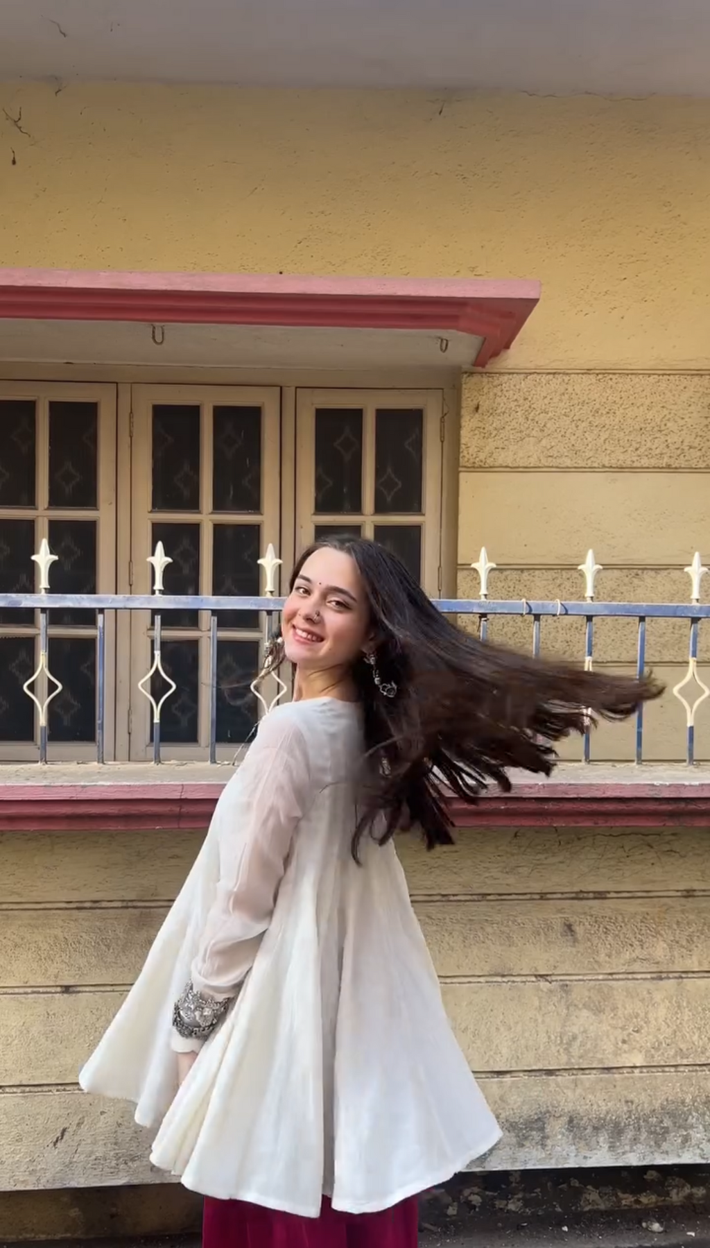 A woman wearing a white long-sleeve tunic top and a deep magenta skirt stands outdoors in front of a yellow building with a window and metal railing, looking back over her shoulder with her hair blowing in the wind.