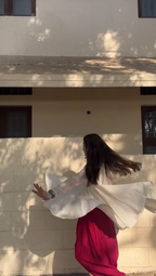 A woman with long dark hair is seen from behind, wearing a flowing white top with wide sleeves and a bright pink skirt, standing in front of a beige stucco wall with dappled sunlight.