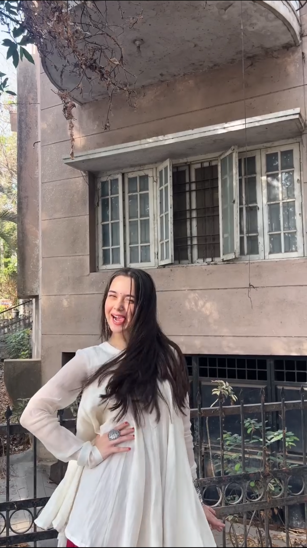 A woman with long dark hair wearing a white long-sleeve dress stands outdoors in front of a weathered beige building with barred windows.