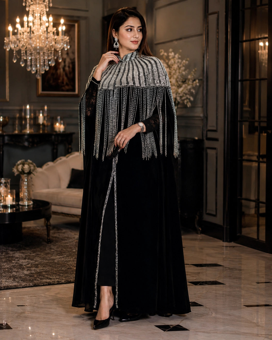 A woman models a luxurious black velvet abaya with a high collar featuring vertical silver beaded fringe, set in an elegant, dimly lit interior with a chandelier and marble floors.