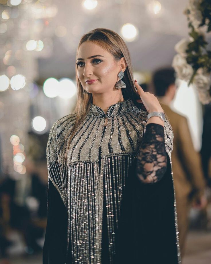 A woman wearing a black and silver embellished high-neck dress with long lace sleeves, photographed in a softly blurred indoor event setting with warm bokeh lights.