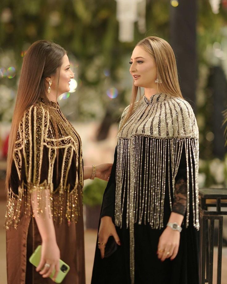 Two women wearing elegant, embellished traditional-style dresses or caftans in an outdoor setting with blurred greenery in the background. The woman on the left wears a dark brown dress with gold metallic fringe and embroidery.