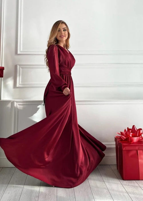 A woman wearing a floor-length burgundy satin gown with long sleeves and a fitted waist, standing in a bright white room with decorative paneling and a red gift box on the floor.