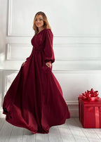 A woman wearing a long-sleeved, floor-length burgundy dress with pockets, standing in a bright room with white paneled walls and light wood floors. A red gift box with a large red bow sits on the floor to her right.