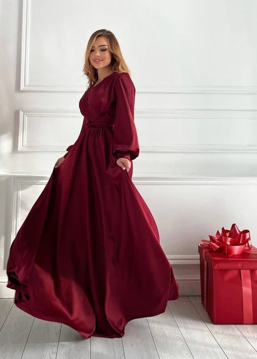 A woman wearing a long-sleeved, floor-length burgundy dress with pockets, standing in a bright room with white paneled walls and light wood floors. A red gift box with a large red bow sits on the floor to her right.