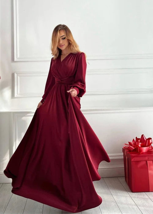 A woman wearing a long-sleeved, floor-length burgundy dress with a wrap-style waist, standing in a bright room with white paneled walls and a light wood floor. A red gift box with a red bow sits on the floor to the right.